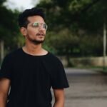 Portrait of a young man wearing glasses, standing outdoors in a park. Casual and relaxed setting.