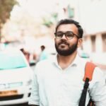 Confident man with glasses and backpack on a sunny urban street with cars in the background.